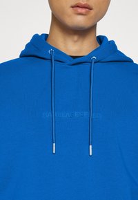 Persona che indossa un hoodie blu brillante con coulisse abbinate e il logo in rilievo "KARL LAGERFELD" sulla parte anteriore.
