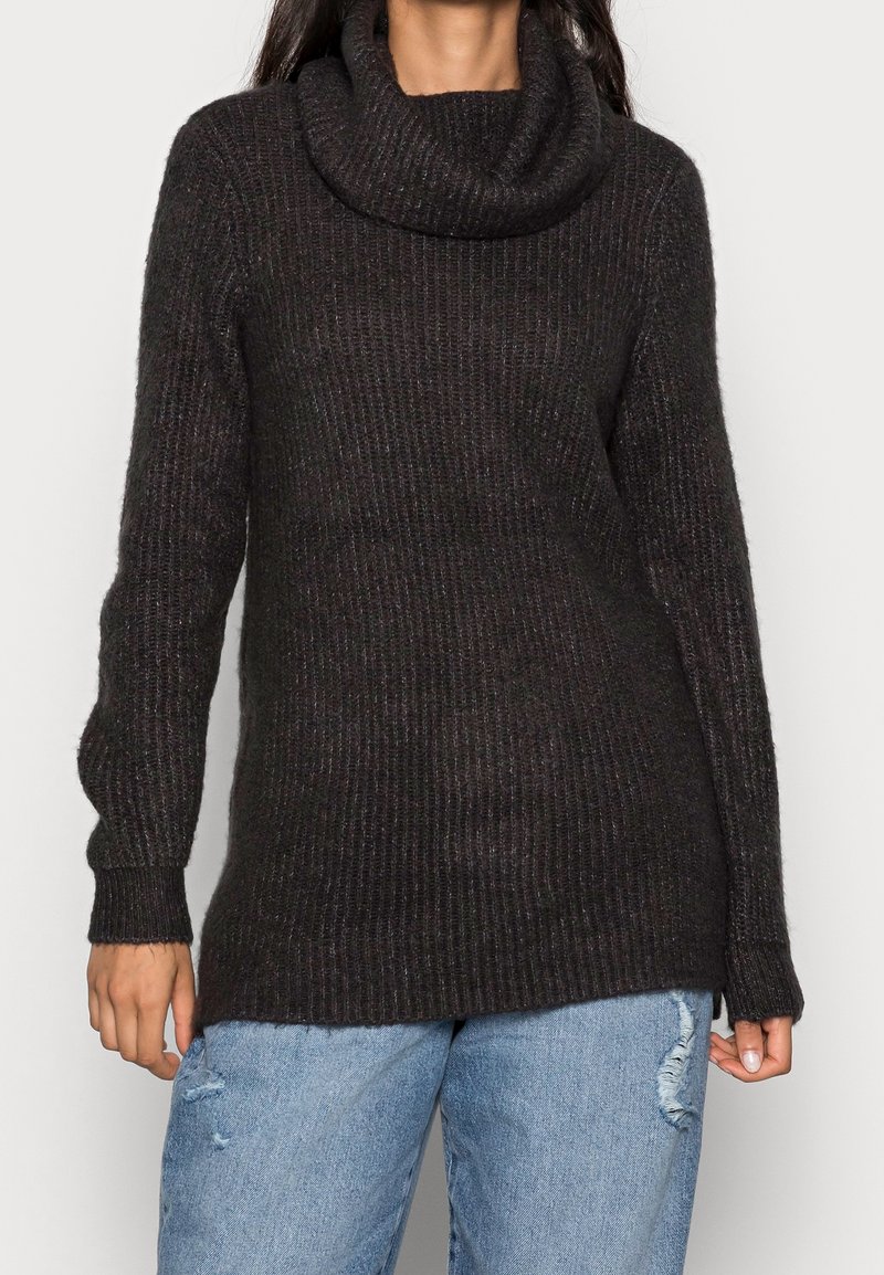 Femme portant un pull à col roulé en maille côtelée noire à manches longues et un jean bleu clair usé.