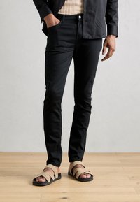 Mustat slim-fit-housut yhdistettynä beigekläisiin korsettikenkiin, joissa on tekstuurilla varustettu hihnapohja ja tasapohja puulattialla.