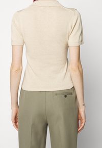 Polo a maniche corte beige con una texture a maglia, dotato di colletto classico e polsini a coste. Indossato con pantaloni su misura verde oliva.
