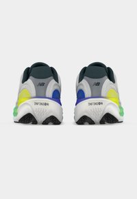 Ζευγάρι γκρι και λευκά παπούτσια τρεξίματος New Balance με μαύρες σόλες, μπλε και κίτρινες λεπτομέρειες, από πίσω, σε λευκό φόντο.