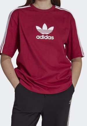 Osoba nosząca czerwoną koszulkę Adidas z białym logo i paskami na rękawach, w połączeniu z czarnymi spodniami Adidas z białymi paskami.