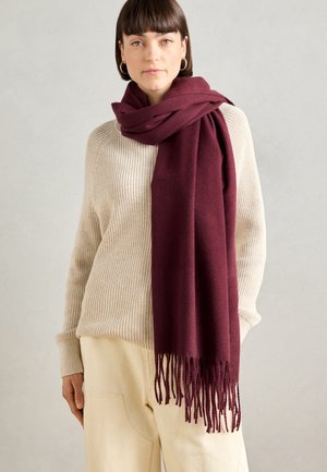 Bordeaux tørklæde lavet af blødt stof, draperet rundt om halsen med frynser i kanten. Båret over en struktureret crèmfarvet sweater.