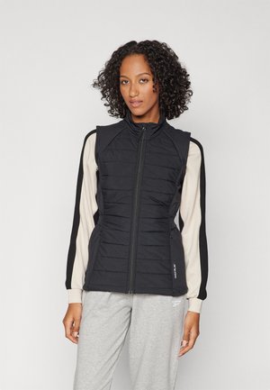 Gilet imbottito nero con colletto alto, design trapuntato verticale, zip frontale e tasche laterali; abbinato a una camicia a maniche lunghe di colore chiaro.