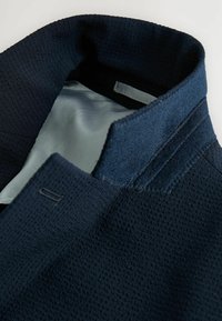 Marineblaue Jacke mit Kragen, der einen strukturierten Stoff und ein glattes Innenfutter zeigt. Verfügt über Schneiderdetails und einen dezenten Glanz.
