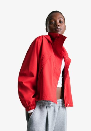 Young woman wearing a bright red jacket, white crop top, and gray sweatpants, posing confidently against a white background.