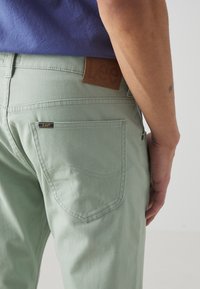 Hombre con pantalones Lee de color verde claro y una camisa azul, mostrando el bolsillo trasero con la etiqueta de la marca Lee y un parche de cuero Lee en la cinturilla.
