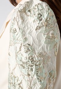 Crème-kleurig kledingstuk met getextureerde mintgroene en gouden bloemmotieven op de mouw. Zachte stof met ingewikkelde details en glans.