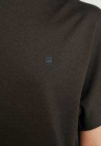 Gros plan d'un t-shirt en tissu maille vert olive foncé avec un petit logo noir G-Star RAW sur le haut de la poitrine et une manche courte visible.
