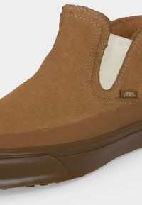 Chaussure marron en daim à enfiler avec panneau latéral élastique, semelle en caoutchouc épaisse et étiquette avec le logo Vans près du col de la cheville.