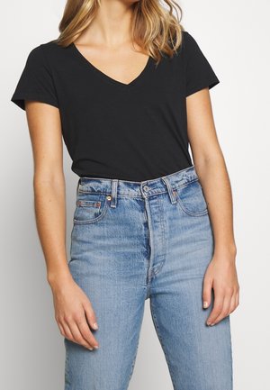 Mujer con camiseta negra de manga corta y cuello en V metida en unos vaqueros de tiro alto azul claro sobre un fondo liso.