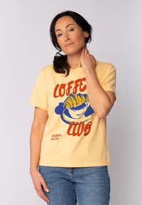 Gelbes Baumwoll-T-Shirt mit "COFFEE CLUB" Grafik, die ein Croissant und eine Kaffeetasse in Blau und Rot zeigt. Kurze Ärmel, entspannte Passform.