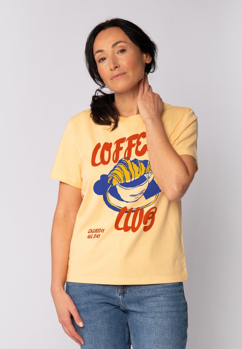 Gelbes Baumwoll-T-Shirt mit "COFFEE CLUB" Grafik, die ein Croissant und eine Kaffeetasse in Blau und Rot zeigt. Kurze Ärmel, entspannte Passform.