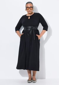 Musta, kolme-neljännes hihainen mekko, jossa on avainrei'ellä varustettu kaula-aukko ja musta nahkavyö vyötäröllä. Taskut kummallakin puolella. Karheat kengät.