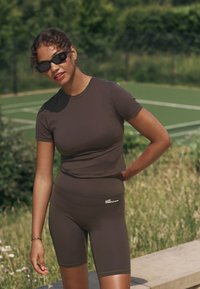 Barna aktív viselet szett, amely egy testhez simuló rövid ujjú felsőt és magas derekú rövidnadrágot tartalmaz. Sima anyagból készült, logó díszítéssel.