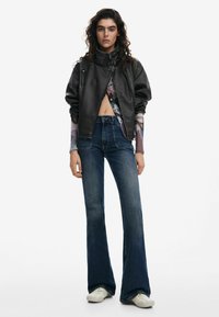 Black leather bomber jacket with high collar, paired with high-waisted flared jeans. Model wears white sneakers; layered graphic top visible.