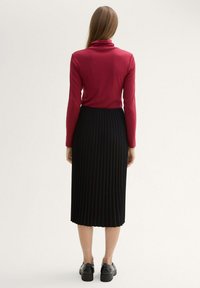 Cremfarbene Langarmbluse mit hohem Kragen, kombiniert mit einem schwarzen, plissierten Midirock; schwarze Schuhe vervollständigen das Outfit aus der Rückansicht.