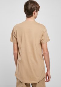 Beige tričko s krátkým rukávem a zaobleným lemem, z pohledu zezadu. Jednoduchý design, bez viditelných log nebo vzorů.