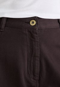 Pantaloni marroni scuri realizzati in tessuto testurizzato, con un bottone dorato e cuciture visibili lungo le tasche e la vita.