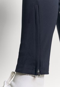 Marina-idrottsbyxor med dragkedja i sidan vid benslutet, tillverkade av ett elastiskt tyg med en slät textur, kombinerade med vita sneakersdetaljer.