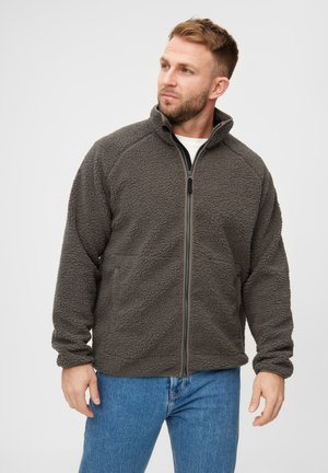 Graue Fleecejacke mit einer strukturierten Oberfläche, voller Reißverschluss und hohem Kragen. Verfügt über Raglanärmel und Seitentaschen. Trägt man zu blauen Jeans.