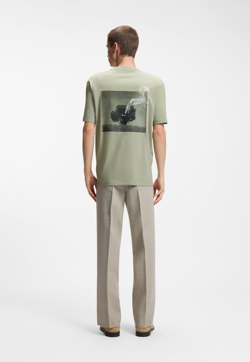 Άνδρας που φοράει ανοιχτό πράσινο μπλουζάκι με μαύρο δέντρο και λευκό πουλί τυπωμένα στην πλάτη, μπεζ παντελόνι και ταμπά παπούτσια, στέκεται με την πλάτη γυρισμένη.