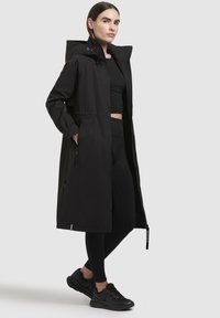 Palton lung negru cu glugă, fermoar frontal și buzunare laterale, asortat cu colanți negri și pantofi sport. Material neted și impermeabil.