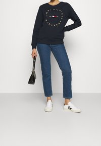 Sudadera navy con letras y logo dorados, combinada con jeans de mezclilla azules y zapatillas blancas con detalles negros, sosteniendo un bolso negro.