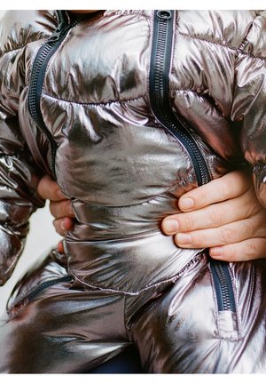 Dorosłe dłonie trzymające dziecko ubrane w błyszczącą srebrną pikowaną kurtkę z czarnymi zamkami, zbliżenie na tułów i dłonie.