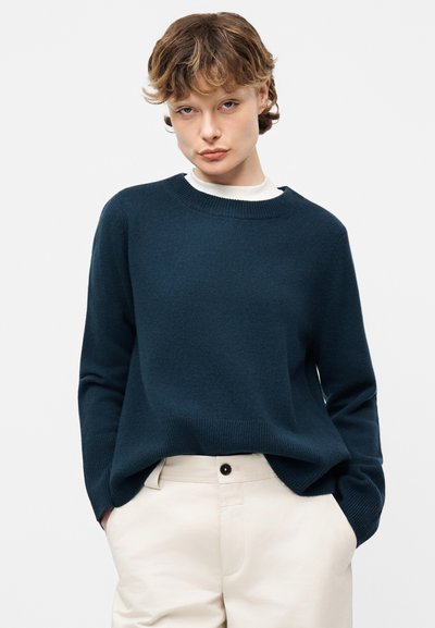 Άτομο με κοντά μαλλιά που φοράει σκούρο μπλε πουλόβερ πάνω από λευκό πουκάμισο και ανοιχτό μπεζ παντελόνι με τα χέρια στις τσέπες, ουδέτερο φόντο.