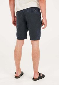 Pantalones cortos azul marino de tejido ligero, con un bolsillo trasero y etiqueta de marca visible. Usados con sandalias negras sobre un fondo neutro.