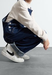 Blå denimoverall med justerbara axelband och metallknappar, i kombination med vita och svarta hög- över sneakers, på ett grått golv.