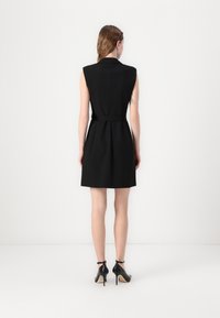Black sleeveless dress with fitted waist, flared skirt, and a smooth texture. Back features a structured design and a belted accent. High-heeled black shoes.
