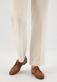Scarpe eleganti in pelle marrone con design perforato, punta rotonda e suola nera, abbinate a pantaloni color crema.