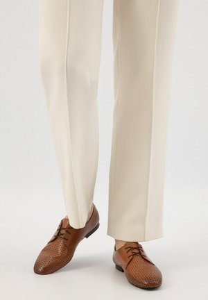 Brown leather dress shoes with perforated design, round toe, and black sole, paired with cream-colored trousers.