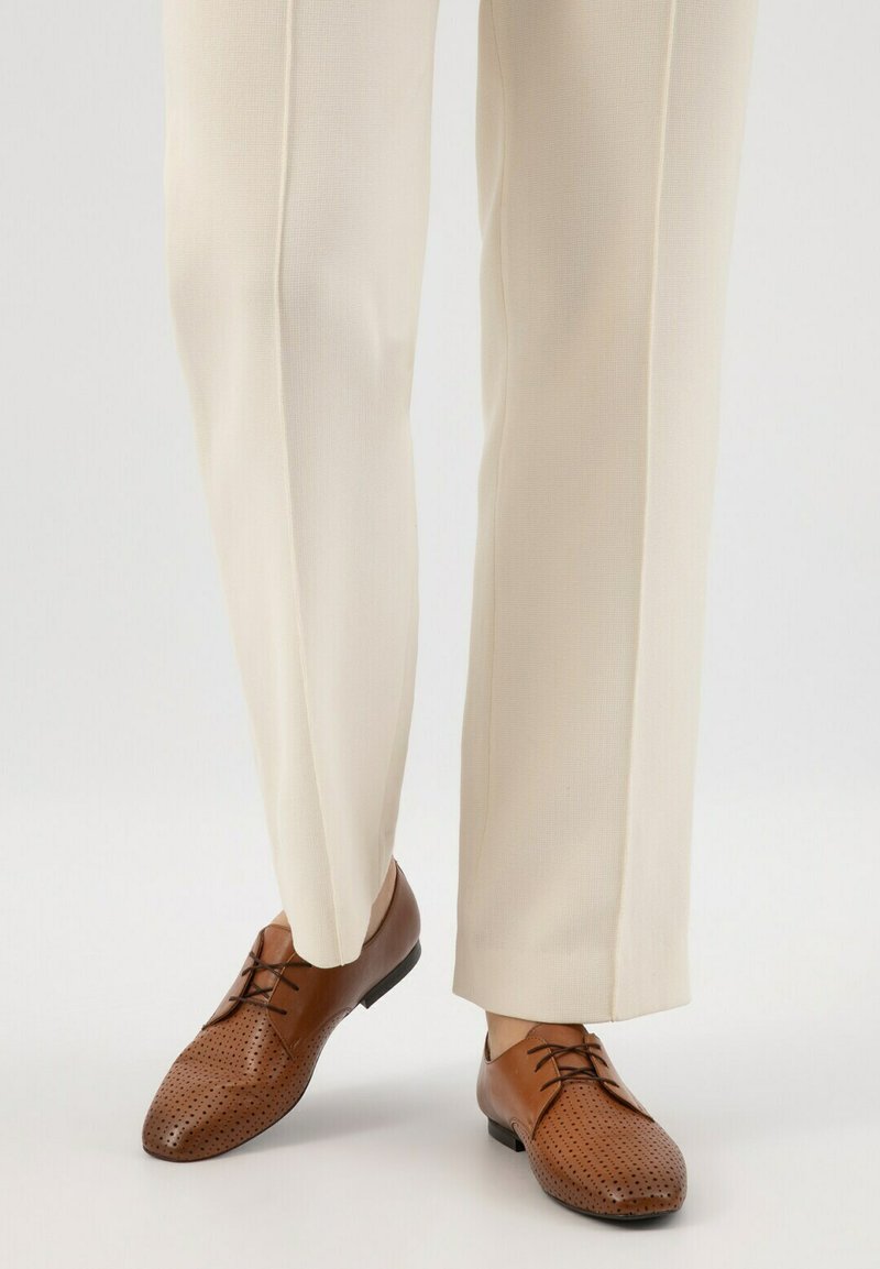 Scarpe eleganti in pelle marrone con design perforato, punta rotonda e suola nera, abbinate a pantaloni color crema.