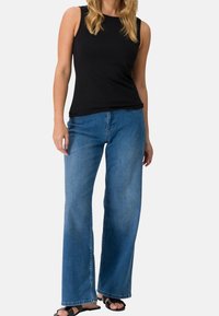 Top nero senza maniche abbinato a jeans a gamba larga blu, con un lavaggio sbiadito e cuciture a contrasto. Il modello indossa sandali neri flat.