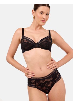 Femme portant un soutien-gorge en dentelle florale noire et une culotte taille haute assortie, posant avec une main sur la hanche devant un fond uni.