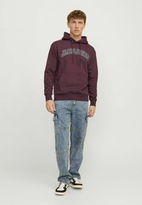 Bordo dukserica s bijelim natpisom "JACK & JONES", džepom s prednje strane, kapuljačom sa vezicom; u paru sa svijetloplavim, zakrpanim cargo trapericama i crnim tenisicama.