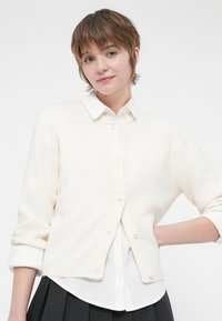 Cardigan bege claro feito de tecido de malha texturizada, com frente de botões, punhos canelados e um corte descontraído sobre uma camisa branca de colarinho.
