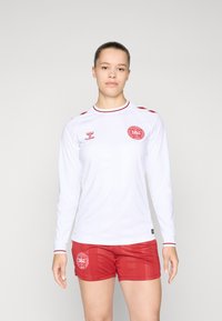 Langærmet hvid fodboldtrøje med røde accenter og et cirkulært logo på venstre bryst, følges af røde shorts med et matchende logo.