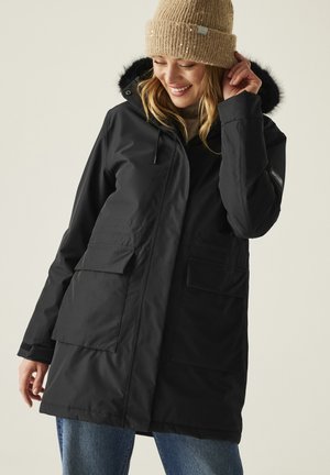 Mujer ajustándose un gorro de punto beige, con un abrigo de invierno negro con capucha forrada de piel y bolsillos con solapa en el frente, sonriendo y mirando hacia abajo.