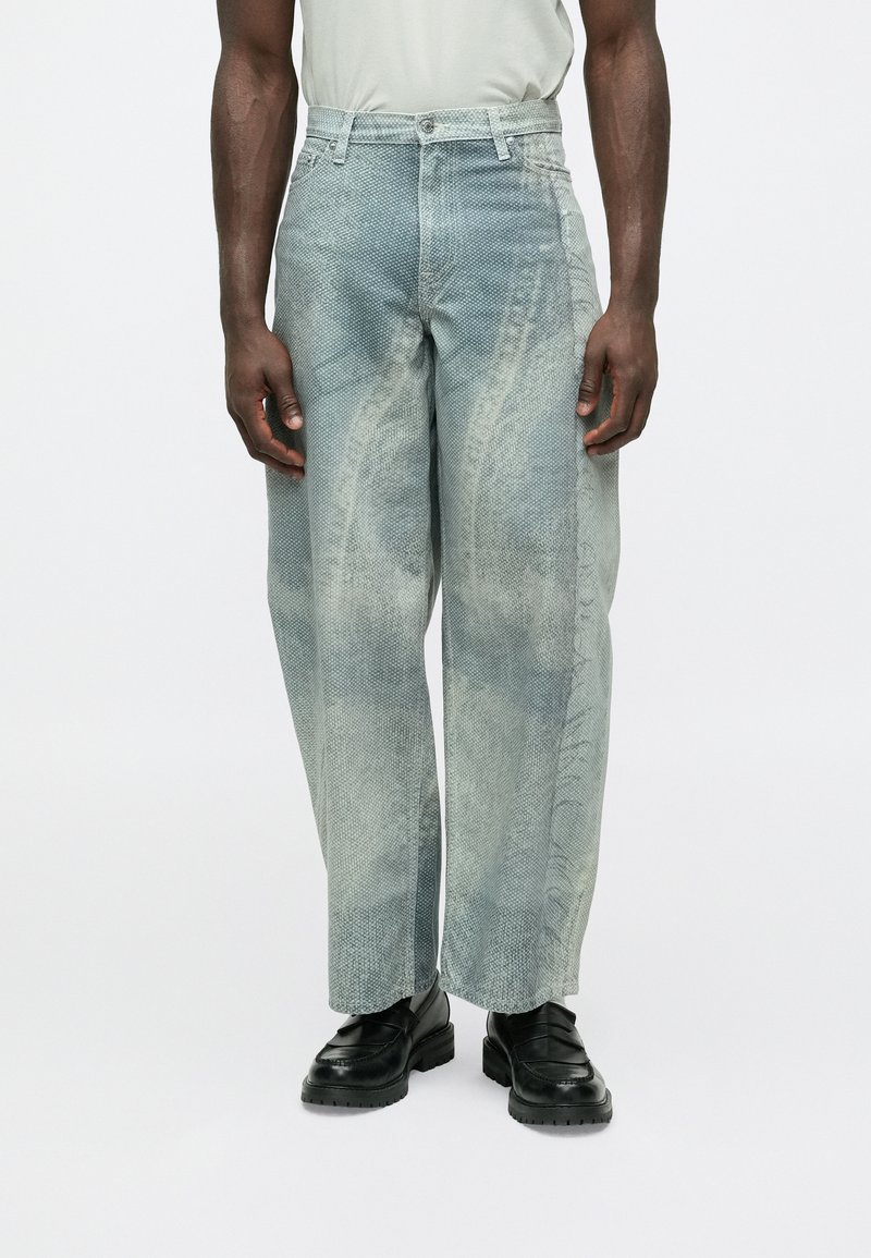 Izblijedjele plave traperice širokog kroja, s teksturiranim završetkom i suptilnim detaljima šavova. Stilizirane s crnom obućom.