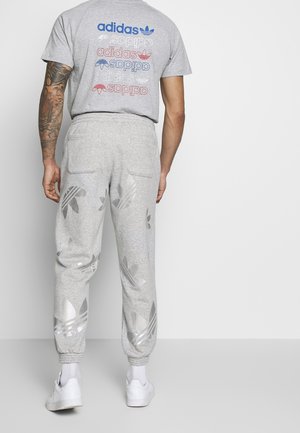 Mężczyzna w szarych dresach Adidas z dużymi wzorami koniczyny oraz w szarej koszulce Adidas z wielokolorowymi logo, stojący na tle białej ściany.