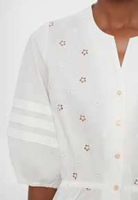 Blouse blanche avec broderie œillet florale, boutons dorés, manches bouffantes avec trois bandes horizontales, portée au niveau du ventre et du bras.