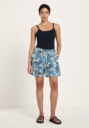 Frau trägt ein schwarzes ärmelloses Oberteil, blaue Shorts mit Paisley-Muster und schwarze Sandalen, steht vor einem schlichten, hellen Hintergrund.