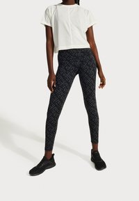 Cropped white t-shirt with short sleeves, paired with black leggings featuring a subtle logo pattern. Black athletic shoes complete the outfit.