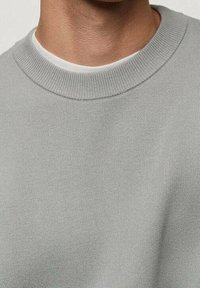 Nahaufnahme einer Person, die einen hellgrauen Rundhals-Pullover über einem weißen Unterhemd trägt, wobei der Hals- und obere Brustbereich sichtbar ist.