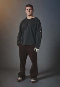 Grå hettegenser med trykknappdetaljer på ermene, kombinert med mørk burgunderbukser og mønstrede sneakers. Ensfarget bakgrunn.