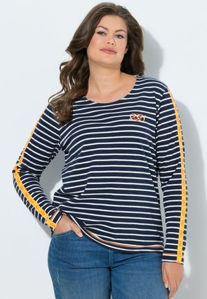 Kvinde iført navyblå og hvidstribet langærmet trøje med orange striber og kringelmønstre ned ad ærmerne, kombineret med blå jeans.